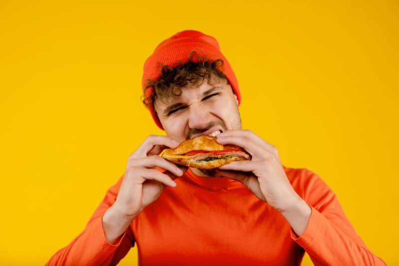Young Handsome Hungry Man Bites Sandwich In Both Hands